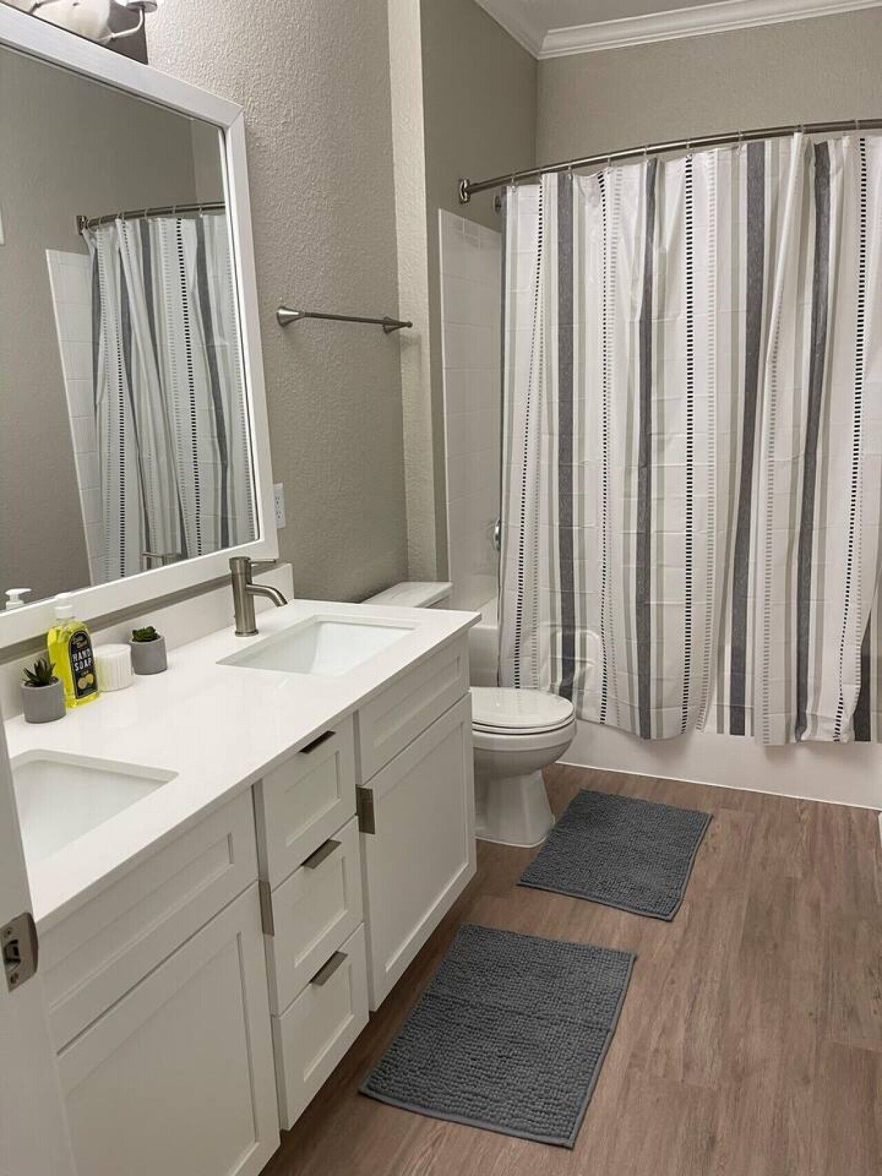 Een badkamer met een wit wastafelmeubel, een grote spiegel, een toilet, een douche met gestreepte gordijnen, en twee grijze vloerkleden.