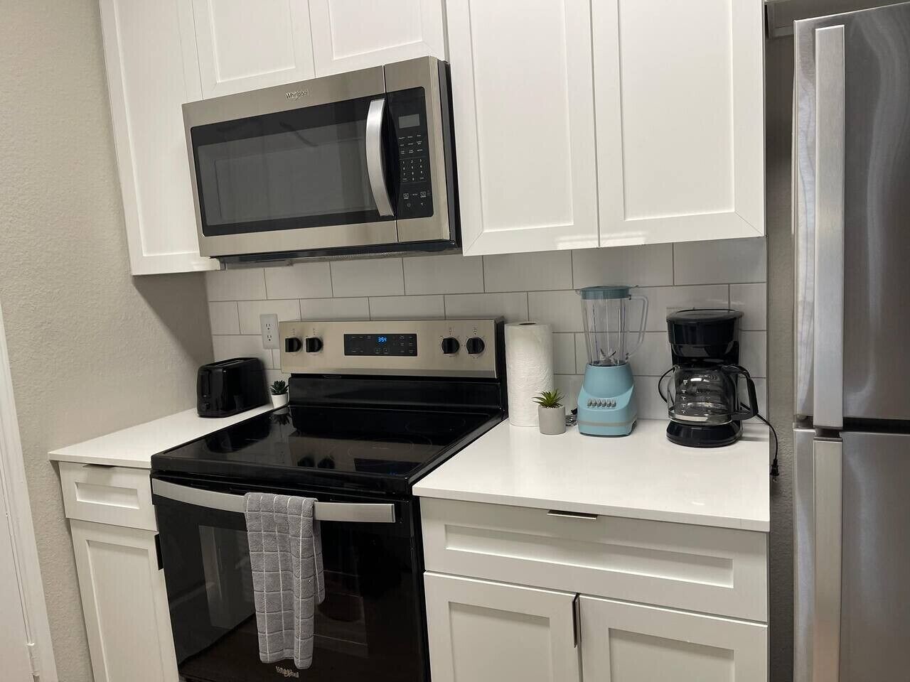 Una cocina con gabinetes blancos, un microondas de acero inoxidable, una estufa negra, una cafetera y un refrigerador.