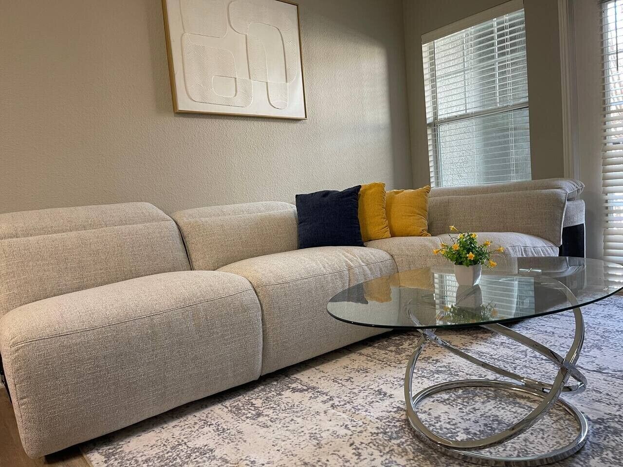 A living room with a beige sofa, a glass coffee table, and a painting on the wall.