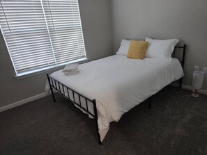 Uma cama de solteiro com estrutura de metal, roupa de cama branca e um travesseiro amarelo. Há uma janela com persianas, piso coberto de carpete e uma parede branca.