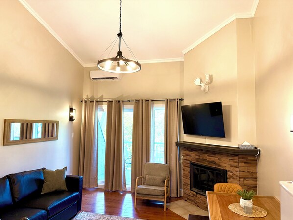 A living room with a sofa, armchair, fireplace, TV, and large windows with curtains. - Nordic Charley - A/C - Mezzanine on 2nd floor - Mountain & lake view (Mont-Tremblant)