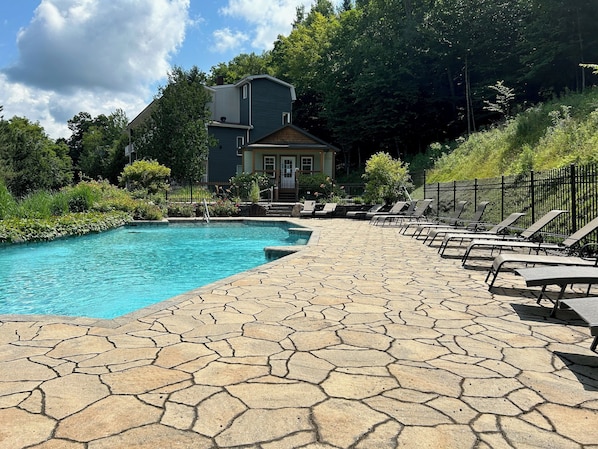 Une piscine avec une terrasse en pierre, des chaises longues et une maison en arrière-plan.
