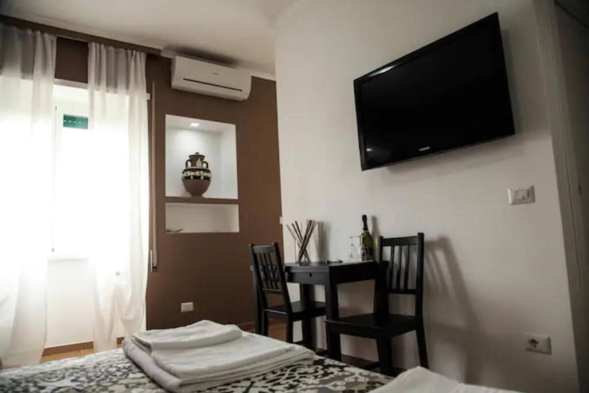 A hotel room with a bed, a TV mounted on the wall, a small table with chairs, and a decorative vase on a shelf.