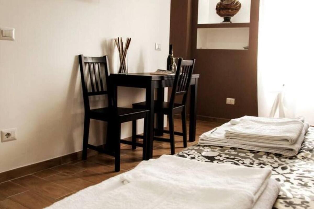 A room with a bed, a table, two chairs, and a small shelf with a decorative item.