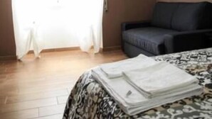 Una habitación con cama, una silla, una mesita, una ventana con cortinas y un estante en la pared con una escultura.