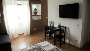 Una habitación con cama, televisor montado en la pared, mesa de comedor con sillas, ventana con cortinas y estante con objetos decorativos.