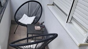 En liten balkong med två svarta fåtöljer i rotting, ett litet bord med en vit kudde och ett fönster med persienner.