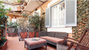 En udendørs terrasse med fletningsmøbler, en polstret bænk og plantekrukker.