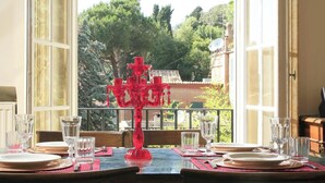 Mesa de comedor preparada para cuatro personas, con vasos transparentes y un centro de mesa de cristal rojo, con vista a un jardín y edificios.