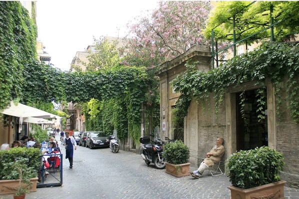 Une rue étroite, bordée de scooters garés, avec des tables installées à l’extérieur et un immeuble à la façade en pierre.
