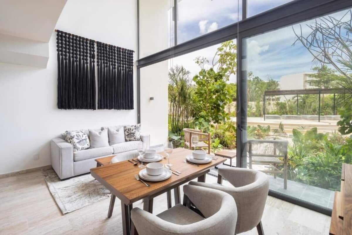 A modern dining area with a wooden table set for four, a grey sofa with patterned cushions, and a large window offering a view of greenery and buildings.