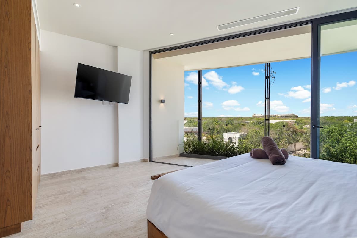 A modern bedroom with a large bed, a flat-screen TV mounted on the wall, and a balcony with a view of the landscape.