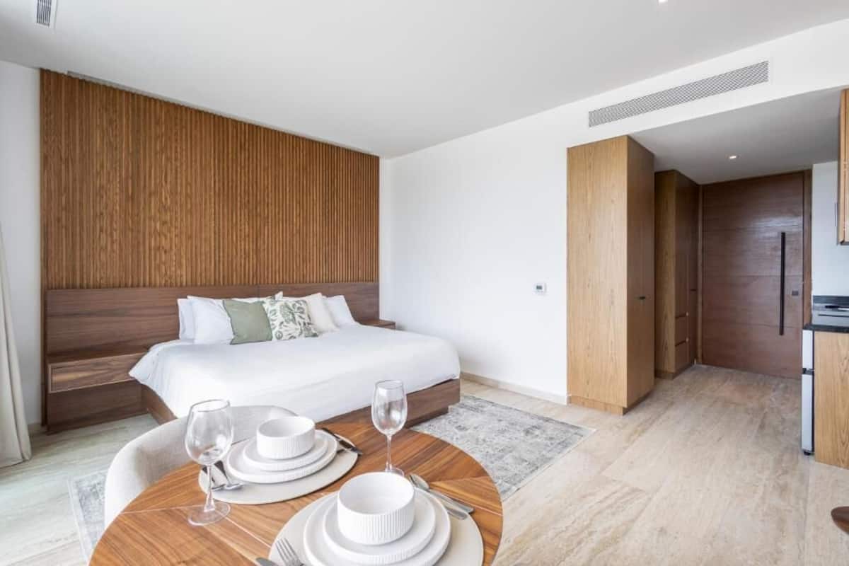 A modern hotel room with a large bed, wooden headboard, a dining table with white tableware, and a kitchenette area.