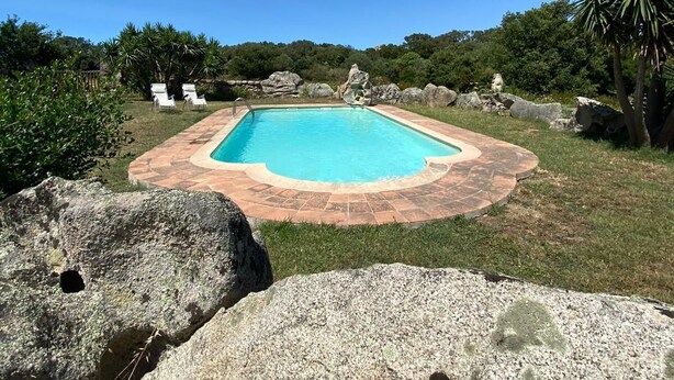 A rectangular pool with a stone border, surrounded by grass and rocks, with a clear blue sky above.