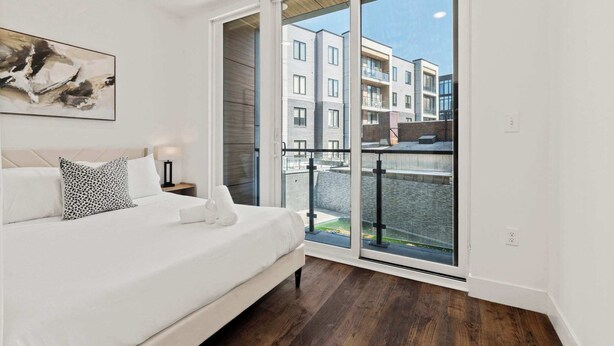 A bedroom with a bed, a nightstand, a lamp, a painting, and a balcony with a view of buildings. - Modern 3BD | Sleeps 8 | Northern Liberties (Philadelphia)