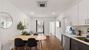 A modern kitchen with white cabinets, a dishwasher, and a sink. There is a dining area with a wooden table and chairs. - Modern 3BD | Sleeps 8 | Northern Liberties (Philadelphia)