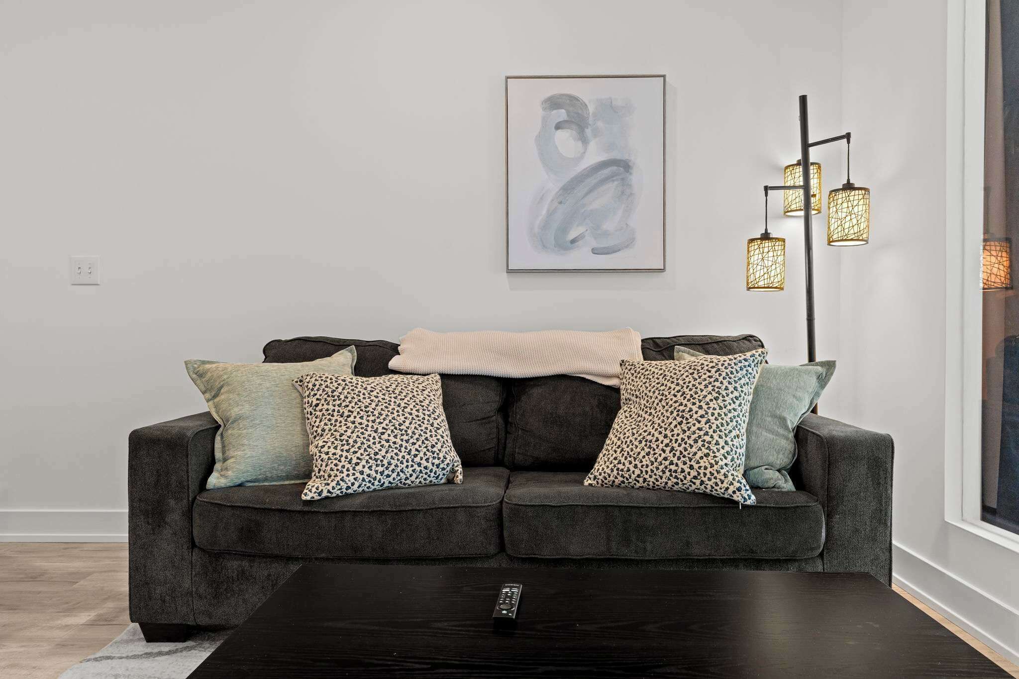 A modern living room with a dark grey sofa, patterned throw pillows, a remote control on a black coffee table, and a framed abstract painting on the wall.