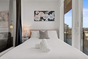 A hotel room with a bed, a painting, a nightstand, a lamp, and a window. - Modern 2BD | Sleeps 6 | Northern Liberties (Philadelphia)