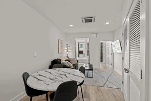 Un salon moderne avec une table en marbre, des chaises noires, un canapé avec des coussins, une petite table d’appoint et un couloir menant à une autre pièce.