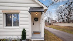 Une maison blanche avec un porche  à l’avant, une boîte aux lettres et un petit arbre.