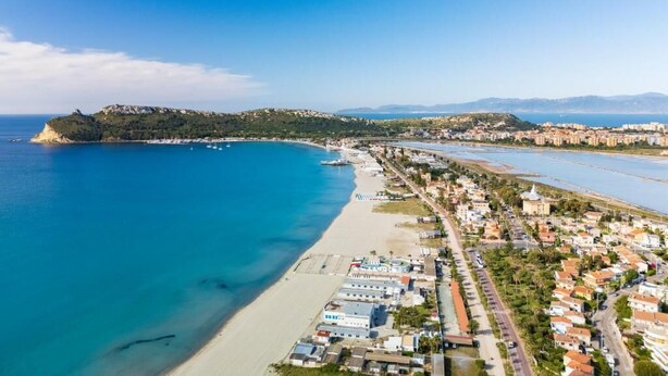Vista aérea de un pueblo costero con una playa, zonas residenciales y una colina destacada al fondo.