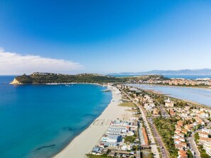 Vue aérienne d’une ville côtière avec une plage, des quartiers résidentiels et une colline imposante en arrière-plan.