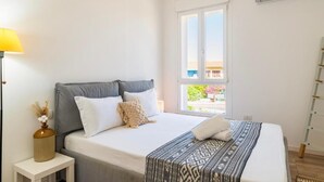 Un dormitorio con cama, mesita de noche con lámpara, ventana con vistas y aire acondicionado.