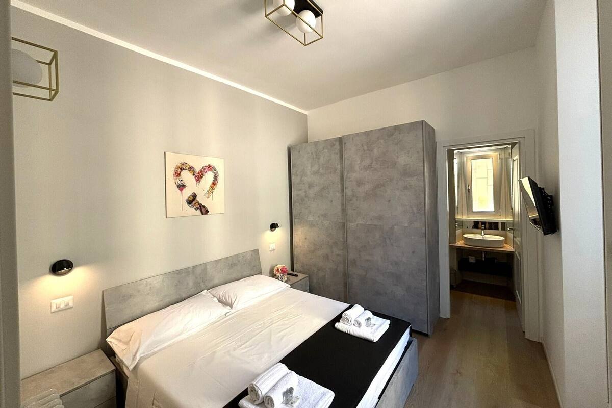 Une chambre moderne avec un lit, des tables de chevet, une œuvre d’art en forme de cœur, une armoire et une salle de bain visible à travers une porte ouverte.