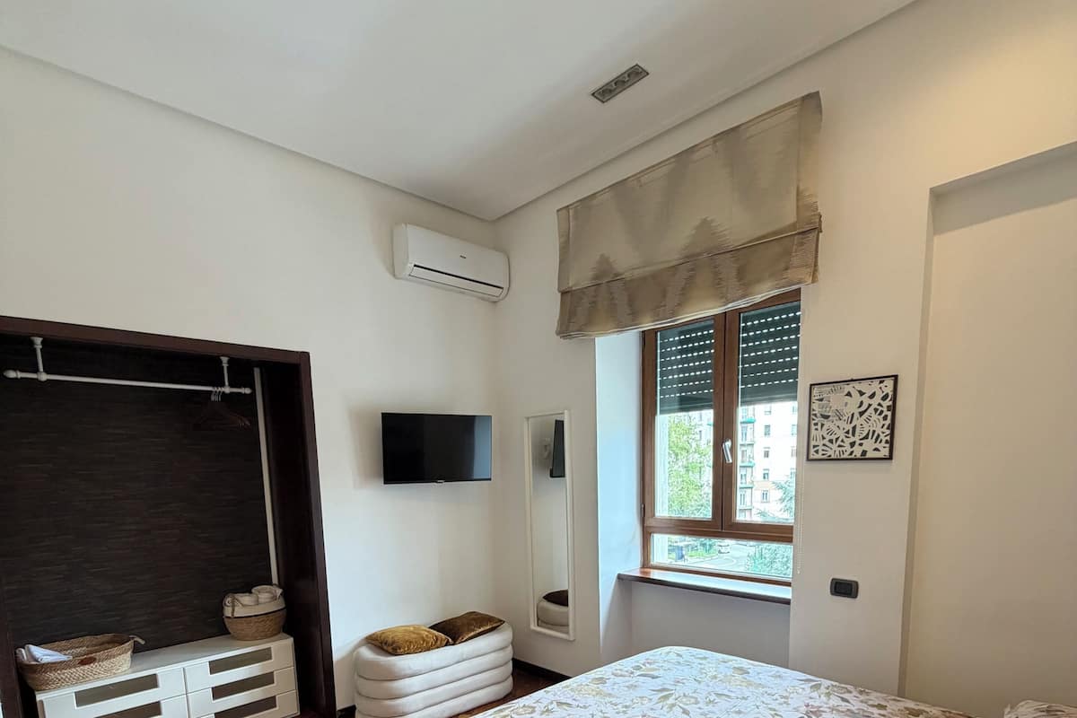 Un dormitorio con cama, televisión, ventana con persianas, armario y mesita de noche.