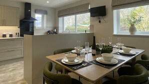 Une cuisine moderne avec un coin repas, un téléviseur fixé au mur et une fenêtre donnant sur les arbres.