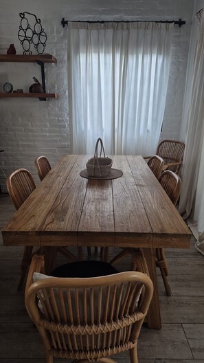 Une table à manger en bois, des chaises en osier, un panier tressé et un rideau blanc en arrière-plan.