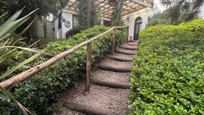 Kivipolku johtaa rakennukseen, jonka päällä on tiilikatto. Rakennuksen ympärillä on vehreää kasvillisuutta ja sen edessä on pergola.