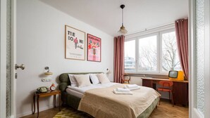 A bedroom with a bed, a desk, a chair, a window with curtains, and two wall-mounted pictures. - Wilcza Retro – City Center, Balcony – by Rentujemy (Warszawa)