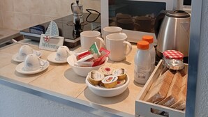 Uma bancada de cozinha com cafeteira, chaleira e micro-ondas. Há pratos, canecas e uma tigela com alimentos.