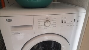 Valkoisen pesukoneen etupuoli, jossa on ohjauspaneeli, sen päällä sinivihreä säiliö ja etiketti, jossa lukee merkki ’beko’.
