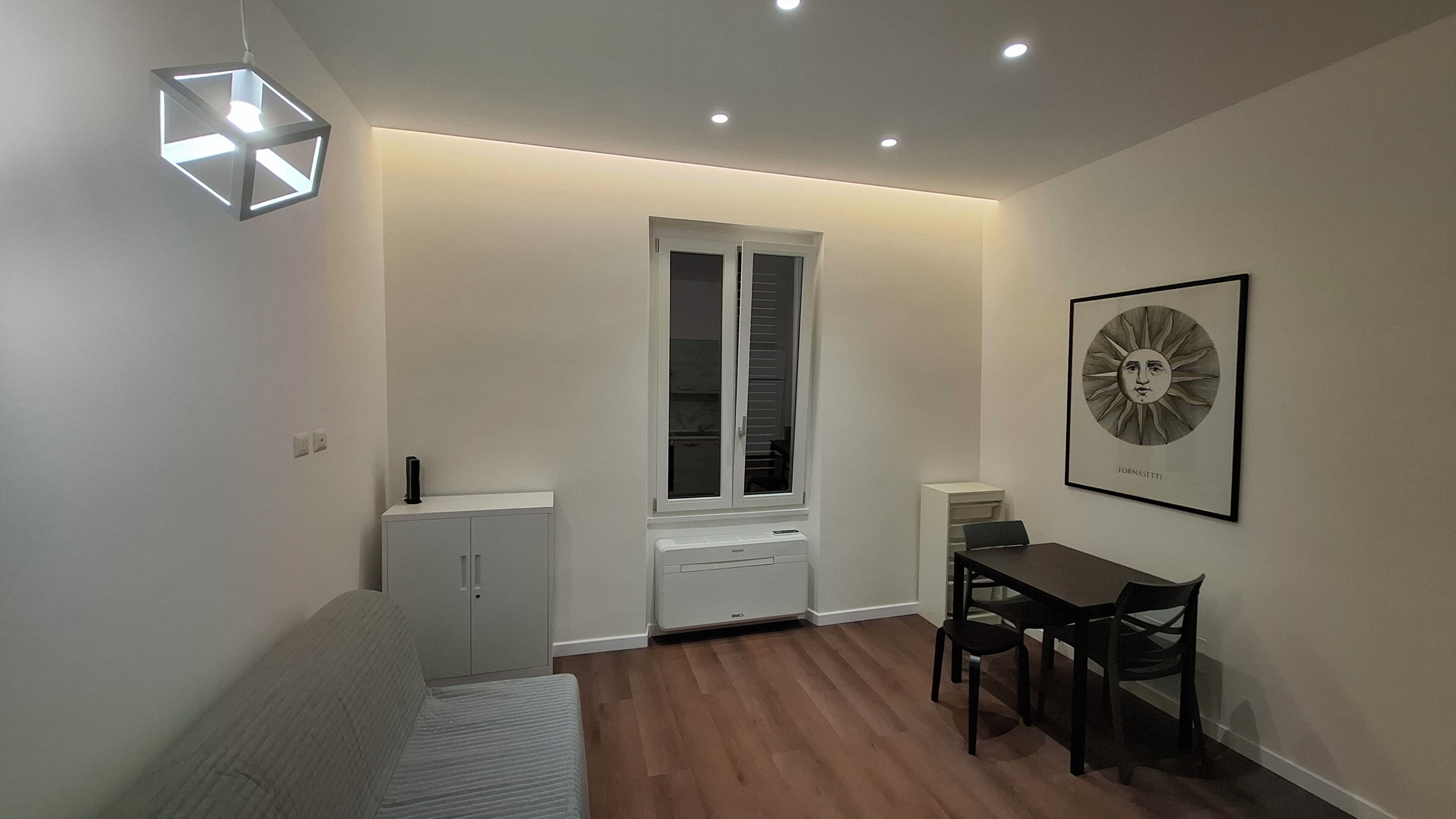 Ein modernes Zimmer mit einer Couch, einem Esstisch mit Stühlen, einem Fenster mit Jalousien, einer Deckenleuchte und einem gerahmten Kunstwerk an der Wand.