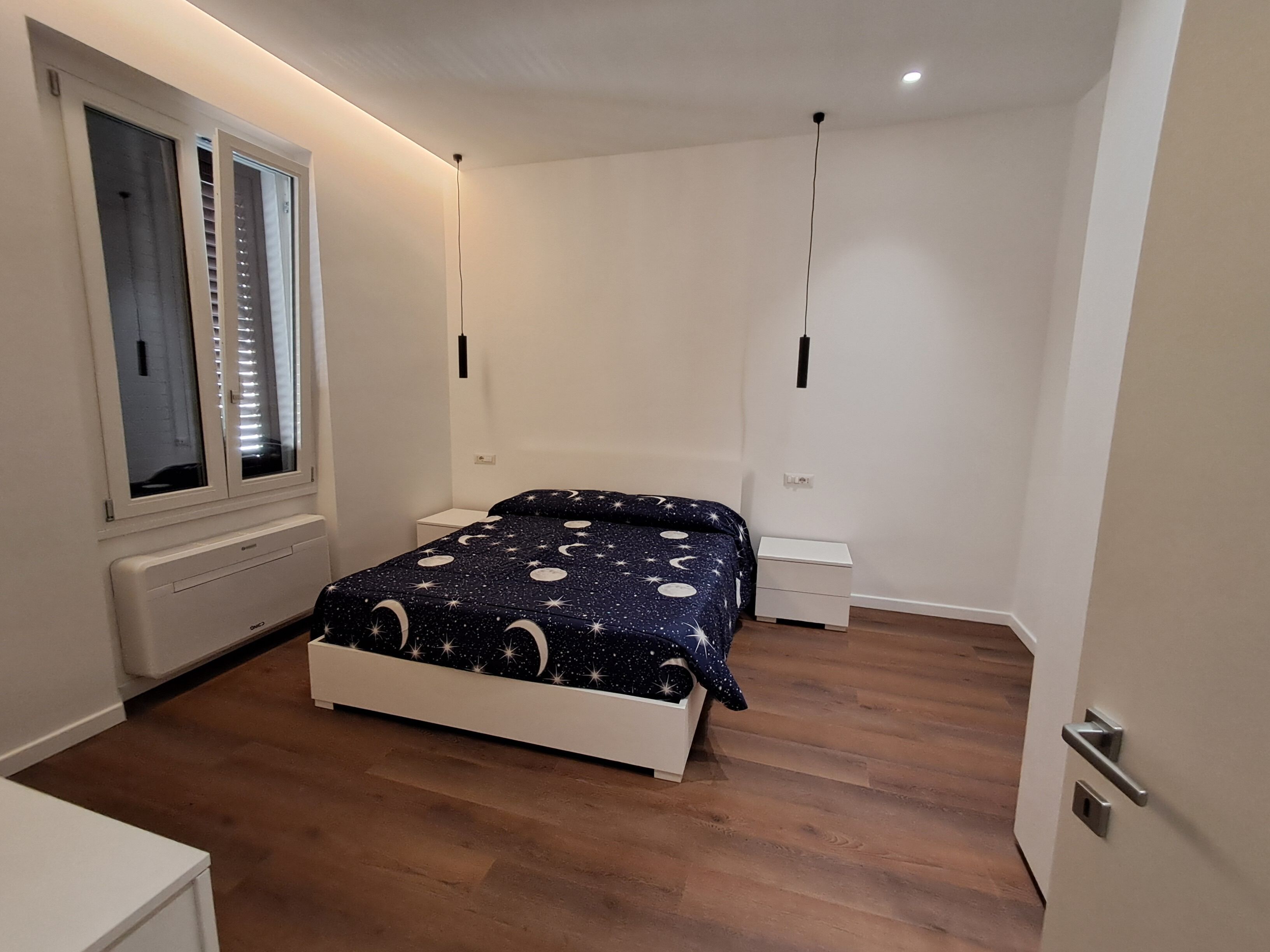 Une chambre à coucher comprenant un lit, une fenêtre avec des stores, une table de chevet et un luminaire fixé au mur.