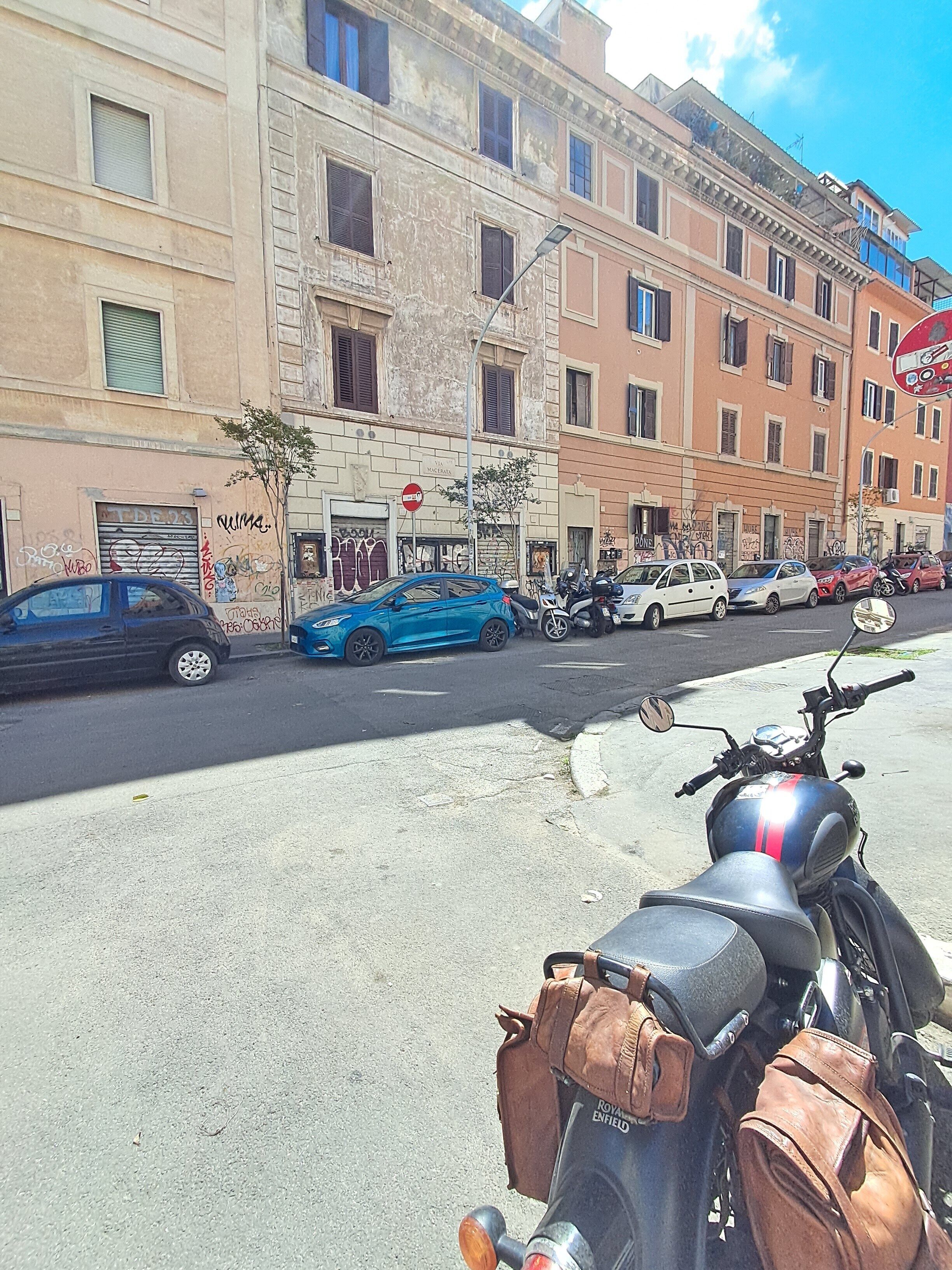 A street view with parked cars, a motorcycle, and buildings with visible signage and windows.