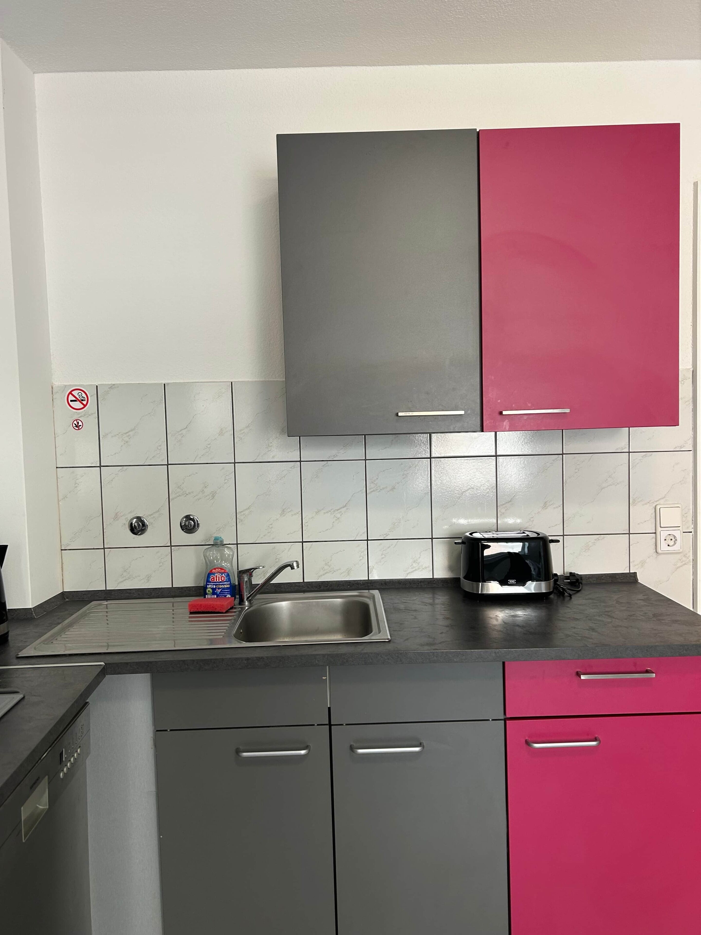 Una cocina con gabinetes grises y rosados, un lavamanos de acero inoxidable y una tostadora en la encimera.