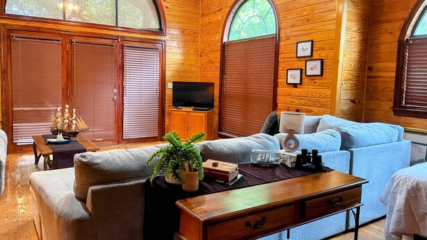 Interior de una cabaña de troncos con sofá, televisor, armarios de madera y ventanas amplias.
