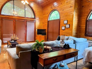 Interior de una cabaña de troncos con sofá, televisor, armarios de madera y ventanas amplias.