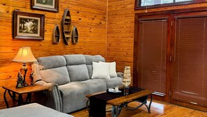 Una habitación acogedora con paredes de madera, un sofá gris, una mesita con una lámpara y un ventanal con persianas.