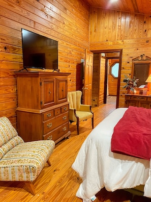 Una habitación con paneles de madera, una cama, un televisor, un tocador, una silla y una puerta que da a otra habitación.