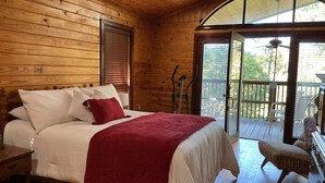 Un dormitorio de cabaña de madera con cama, almohadas, una colcha roja, una mesita de noche, una silla y un balcón con vistas.