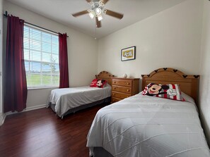 Ein Schlafzimmer mit zwei Betten, einem Deckenventilator, einem Fenster mit Vorhängen und einem Mickey Mouse Kissen.