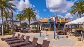Un espace aménagé au bord de la piscine, avec des chaises longues, un bar-restaurant et des palmiers.