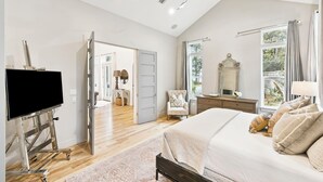 Un dormitorio con cama, televisor sobre un soporte, ventilador de techo, ventana con cortinas y puerta que da a otra habitación.