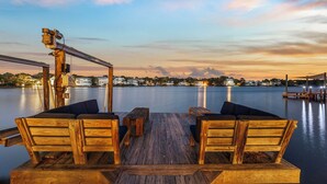 Una terraza de madera con dos bancos y una mesa, con vista a una masa de agua tranquila y edificios al fondo.