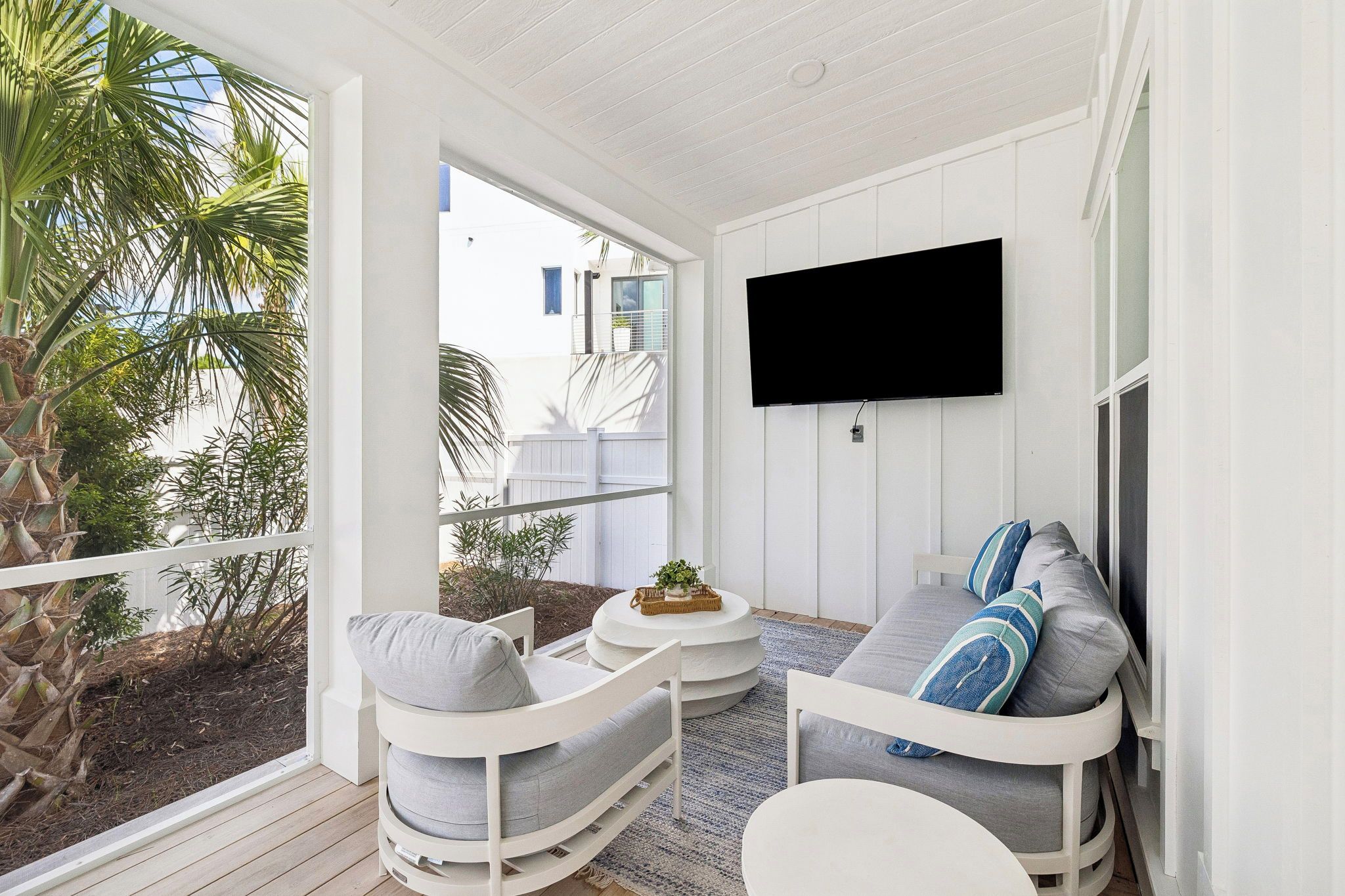 A sunlit patio with a white ceiling, a flat-screen TV mounted on the wall, a grey sofa with blue pillows, and a white chair.