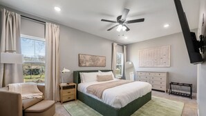 Une chambre à coucher comprenant un lit, des tables de chevet, une chaise, un bureau, un ventilateur de plafond et une fenêtre avec des rideaux.